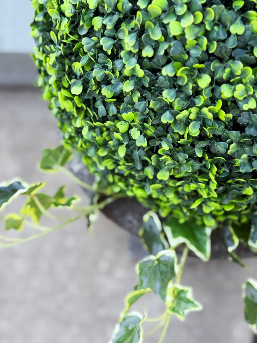 7 easy front porch decor ideas for spring 7 a close-up of a dense, artificial green topiary ball in a planter, with a few ivy leaves and stems extending outward against a blurred gray background.
