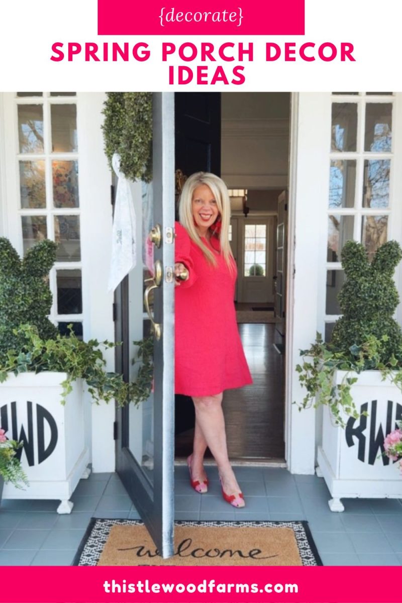 7 easy front porch decor ideas for spring 14 a smiling woman in a bright pink dress stands in a doorway with spring front porch decor ideas, including topiary plants and white planters with black letters. a welcome mat is at her feet, surrounded by charming pink accents.