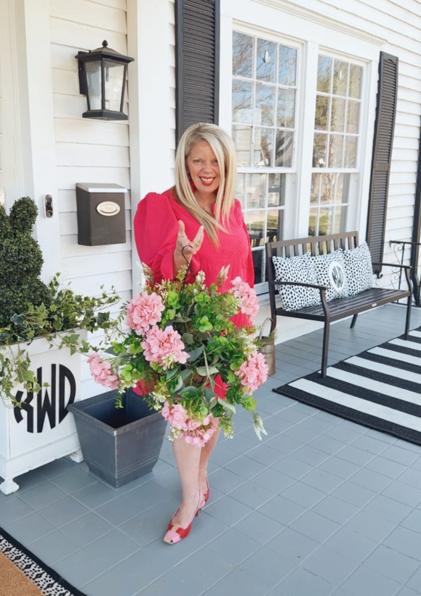7 easy front porch decor ideas for spring 13 woman with long blonde hair wearing a bright red dress and matching shoes stands on a porch holding a large bouquet of pink flowers. the porch is decorated with black and white accents and a bench.