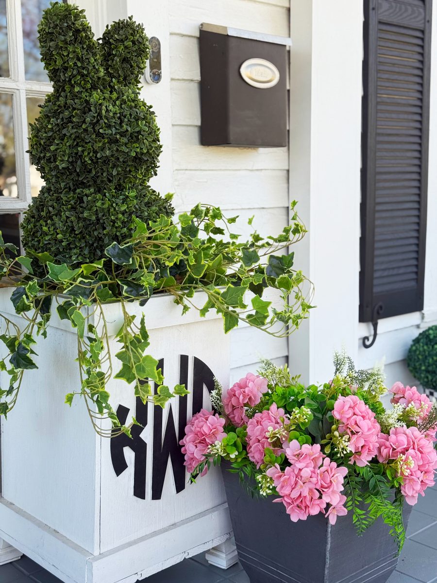 7 easy front porch decor ideas for spring 12 a green topiary shaped like a bunny sits in a white planter with trailing ivy, next to a gray planter filled with pink flowers, outside a white house with black shutters and a black mailbox.
