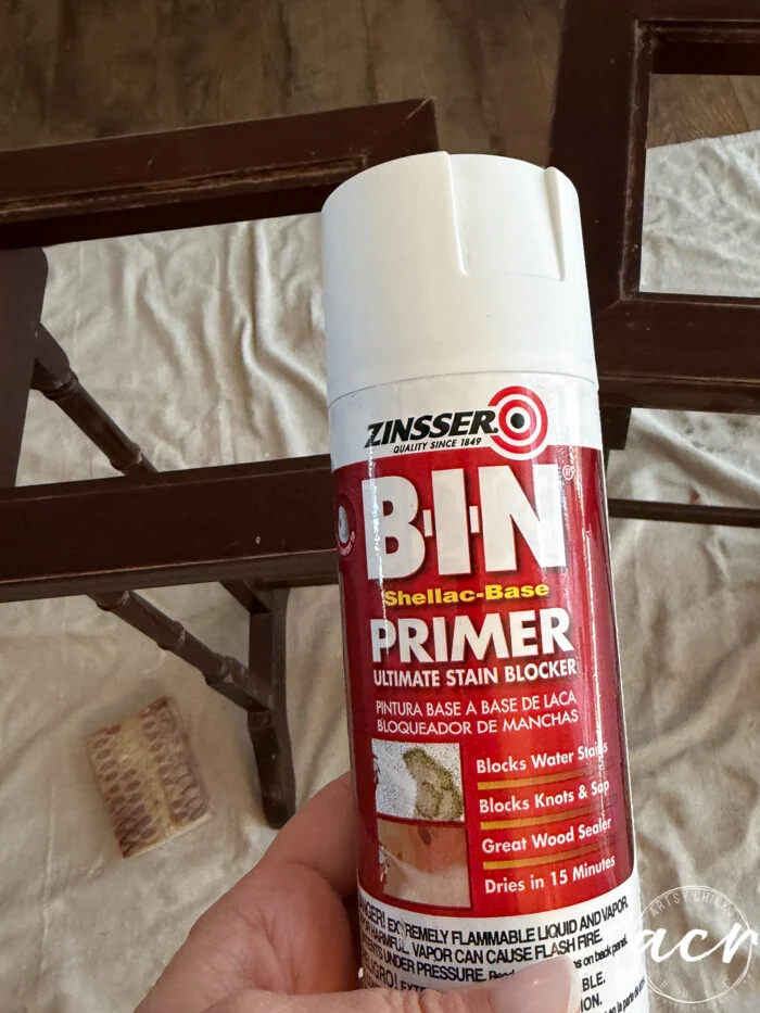 a hand holds a can of zinsser b-i-n shellac-base primer in front of a wooden frame, with a sanding sponge and brown paper on a light surface—perfect for refinishing your glass topped nesting tables.