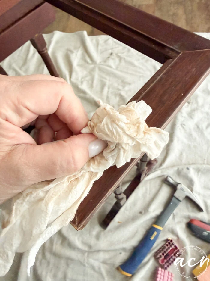 a hand uses a crumpled paper towel to clean a wooden nesting table leg; tools including a hammer and pliers are visible on a cloth-covered floor in the background.