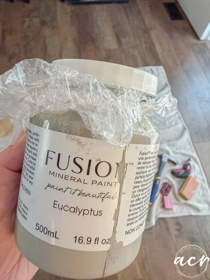 a hand holds a 500ml jar of fusion mineral paint in eucalyptus, with plastic wrap under the lid. paint supplies and a drop cloth surround glass topped nesting tables on a wooden floor in the background.