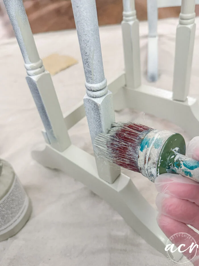 a hand wearing a glove holds a paintbrush, applying white paint to the leg and frame of a wooden chair. nearby, glass topped nesting tables sit on a protective cloth, keeping the workspace tidy.