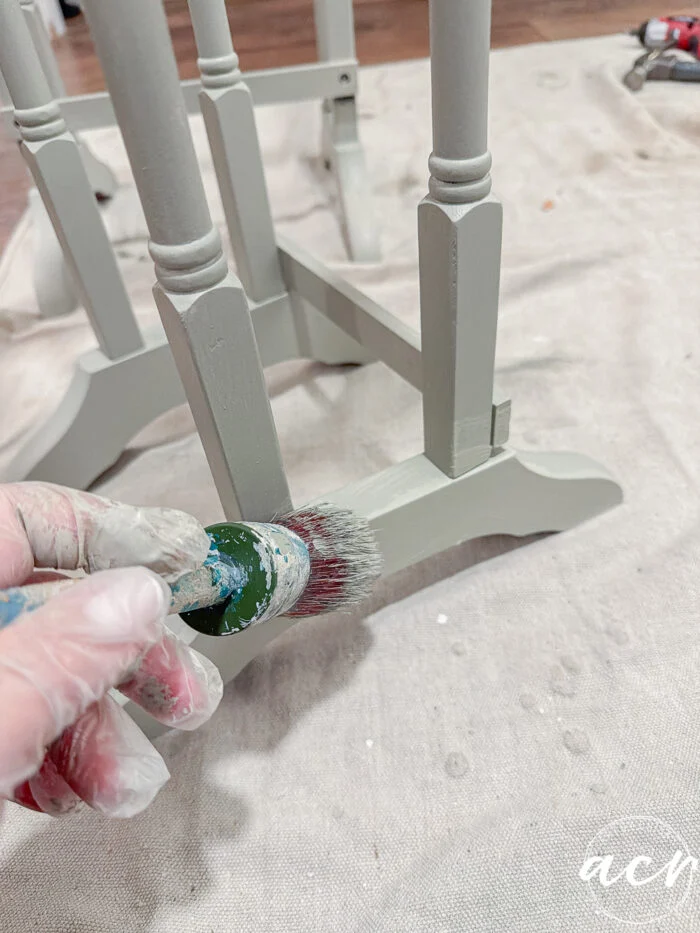 a gloved hand uses a round paintbrush to apply light green paint to the leg and support of a wooden table. glass topped nesting tables and other painting supplies are visible on a protective drop cloth in the background.