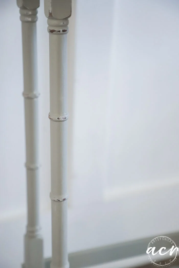 close-up of two light-colored, painted wooden table legs with a bamboo-like design from stylish glass topped nesting tables, set against a plain white background. the bottom right corner features a circular watermark with the letters acv.