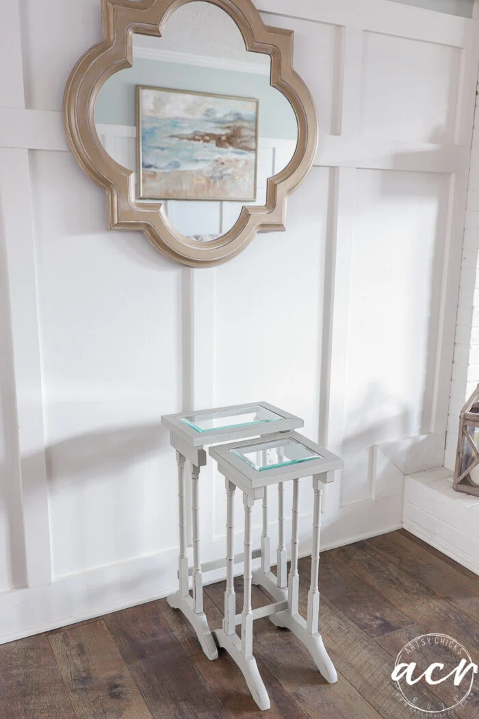 a pair of white glass nesting tables sit against a white paneled wall, beneath a decorative mirror and a framed landscape painting. the floor is dark wood.