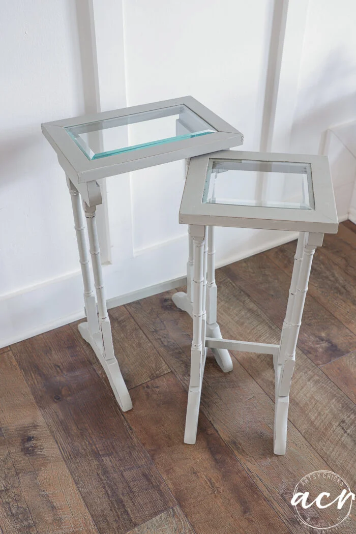 two small, light gray nesting tables with glass tops and faux bamboo-style legs stand on a wooden floor against a white wall. the glass topped tables are different heights and overlap slightly for a chic table update.