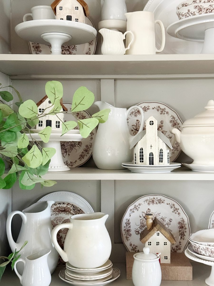 7 simple steps to style a hutch 37 shelves display white and patterned dishes, teapots, pitchers, small ceramic house ornaments, and green foliage. style a hutch for a neat and cozy farmhouse or cottagecore aesthetic with these charming decorative touches.