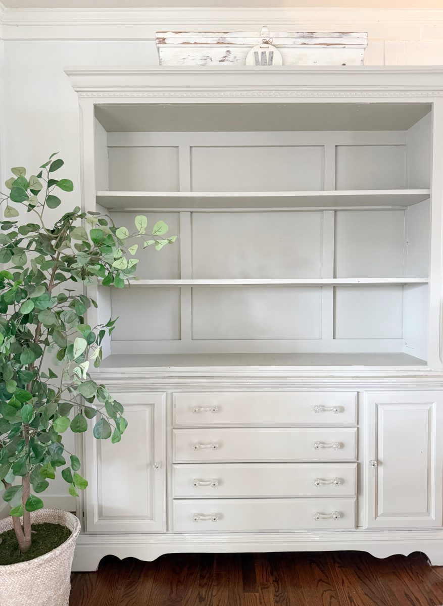 7 simple steps to style a hutch 33 a large white cabinet with empty shelves and drawers stands against a white wall. a potted green leafy plant is to the left, and a vintage white decorative box sits on top—an inviting spot to style a hutch on wooden floors.