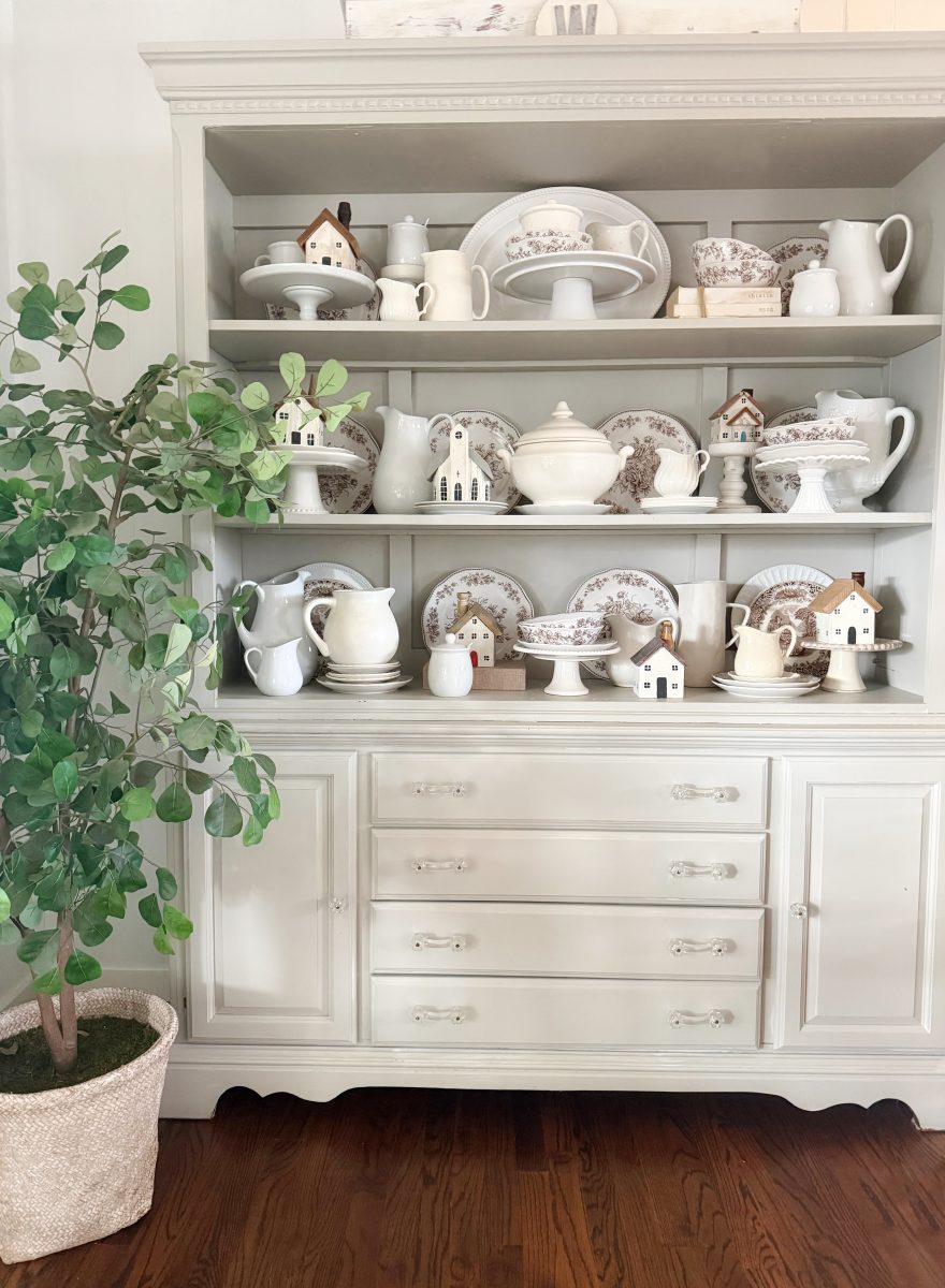 7 simple steps to style a hutch 40 a white china cabinet styled like a hutch displays vintage dishes, pitchers, and tureens in neutral tones, accented with small decorative house figurines. a potted green plant stands to the left on a wooden floor.
