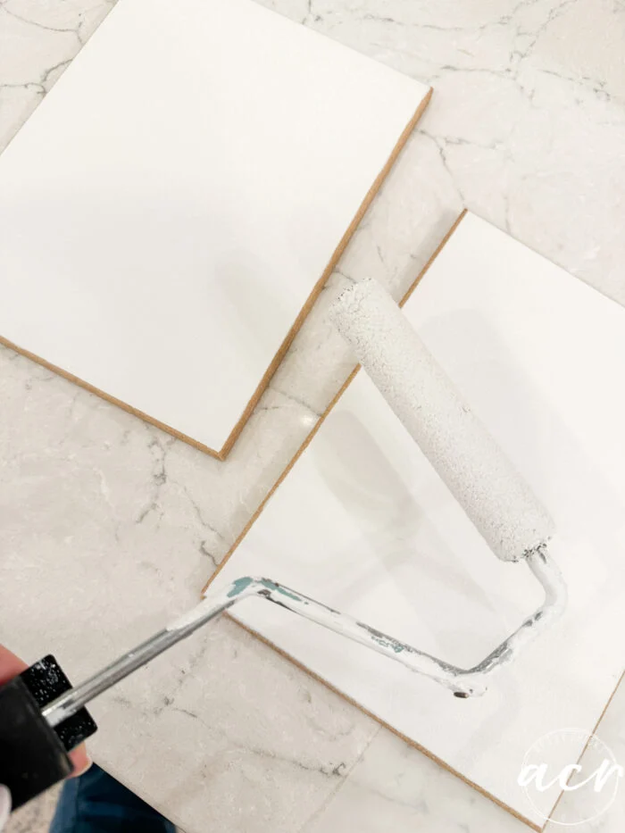 a hand holds a paint roller applying white paint to a blank canvas on a marble surface, with another blank canvas nearby—perfect for your next flipping smalls: easter edition creative project.