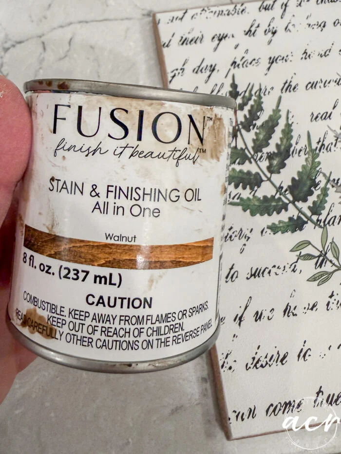 a hand holds a can of fusion stain & finishing oil (walnut, 8 fl. oz.) in front of a decorative art piece with green botanical illustrations and script text—perfect for flipping smalls or creating an easter edition masterpiece. the can displays cautionary information.