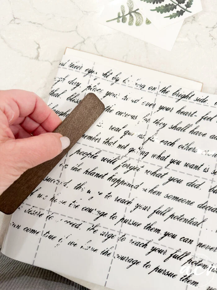 a hand uses a brown tool to rub black script lettering onto a white sheet, with easter edition plant stickers nearby on a marble surface.