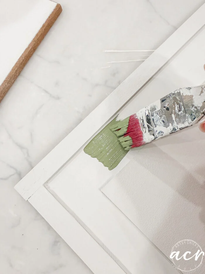 a hand uses a paintbrush to apply light green paint to the white wooden frame of a canvas, on a marble surface—perfect for your easter edition of flipping smalls projects.