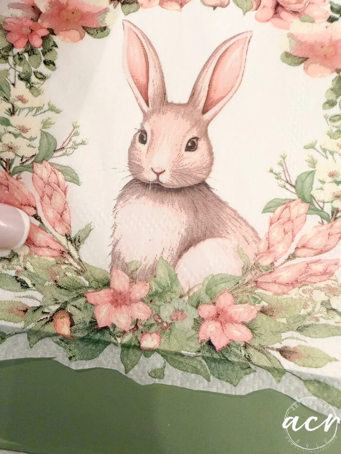 illustration of a brown rabbit with pink ears sitting among pink flowers and green leaves, framed in a floral wreath on a green background. a hand with light-colored nail polish is visible on the left side—perfect for an easter edition of flipping smalls.
