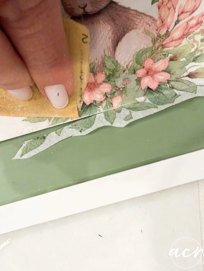 a hand with light nail polish uses sandpaper to smooth the edge of a floral and bunny illustration on a green and white surface, creating an adorable flipping smalls piece for the easter edition.