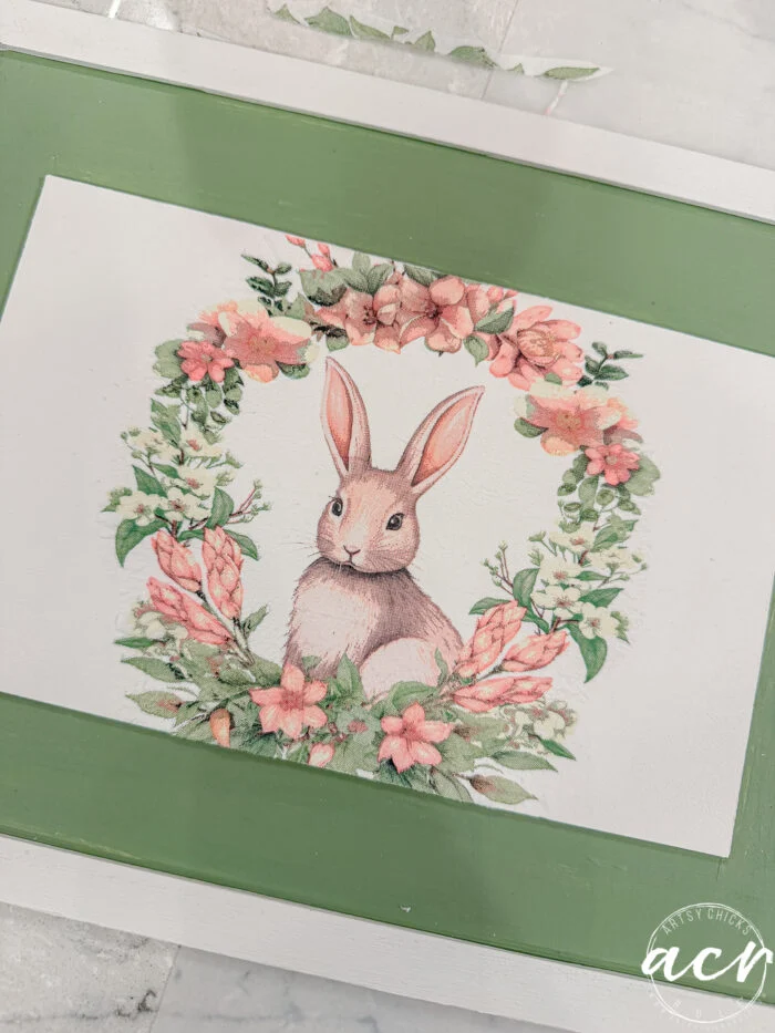 a painted illustration of a brown bunny sitting inside a wreath of pink and green flowers, bordered by a green rectangular frame. this easter edition image has a soft, pastel aesthetic perfect for flipping smalls collections.