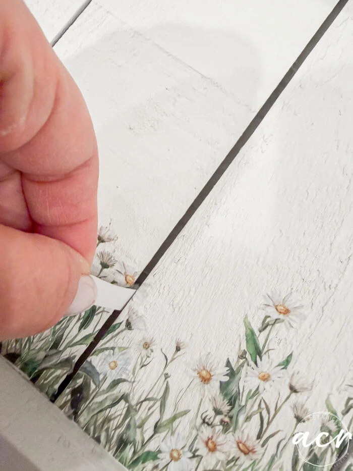 a hand peels up a strip of decorative floral transfer from a white wooden surface, revealing a painted design of green leaves and small white flowers—perfect for your flipping smalls easter edition craft projects.