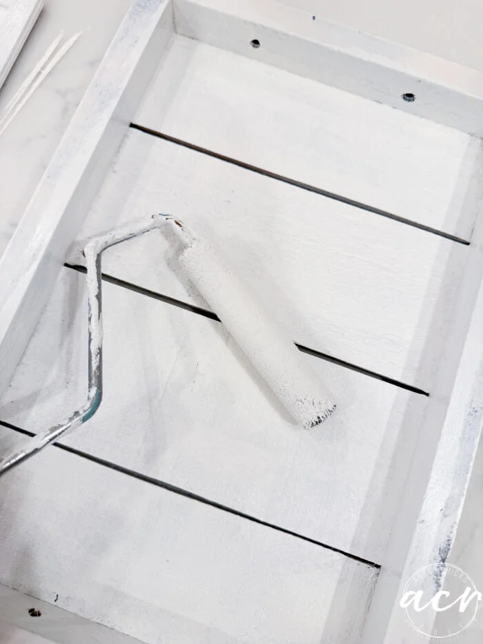 a small paint roller covered in white paint rests inside a white wooden tray with slats. the tray appears freshly painted, adding a clean, bright touch—perfect for a flipping smalls easter edition refresh.