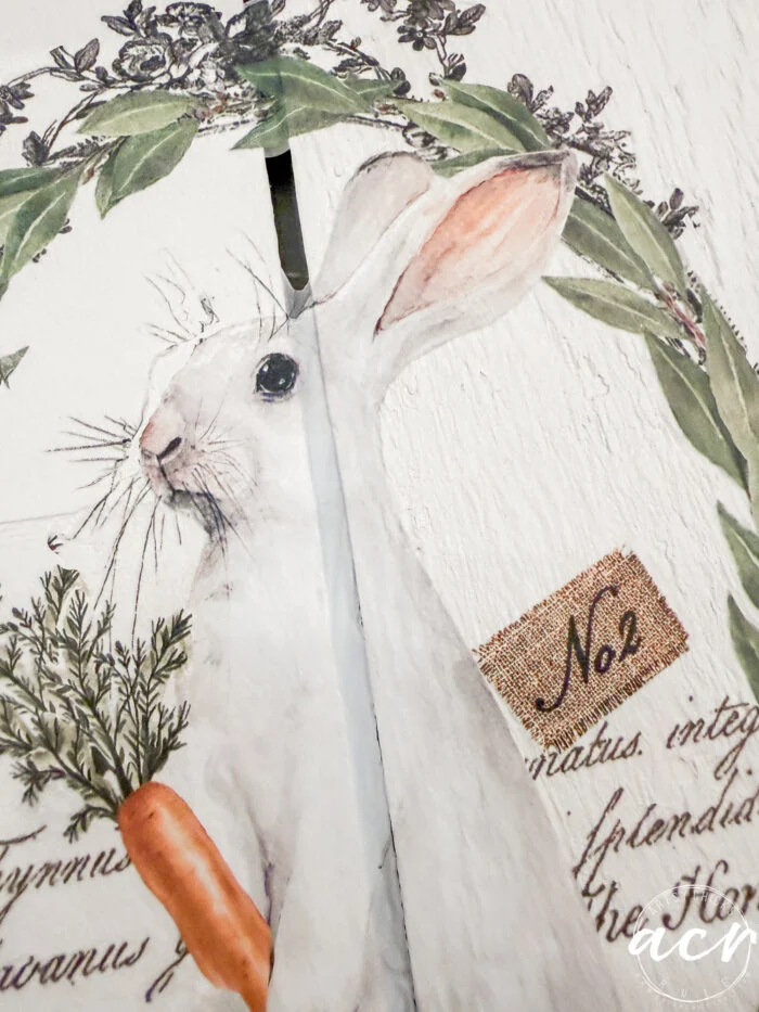 a painted illustration of a white rabbit holding a carrot is partially split by a wooden seam. the background features green foliage, cursive script, and a small "easter edition" label with no 2 written on it.