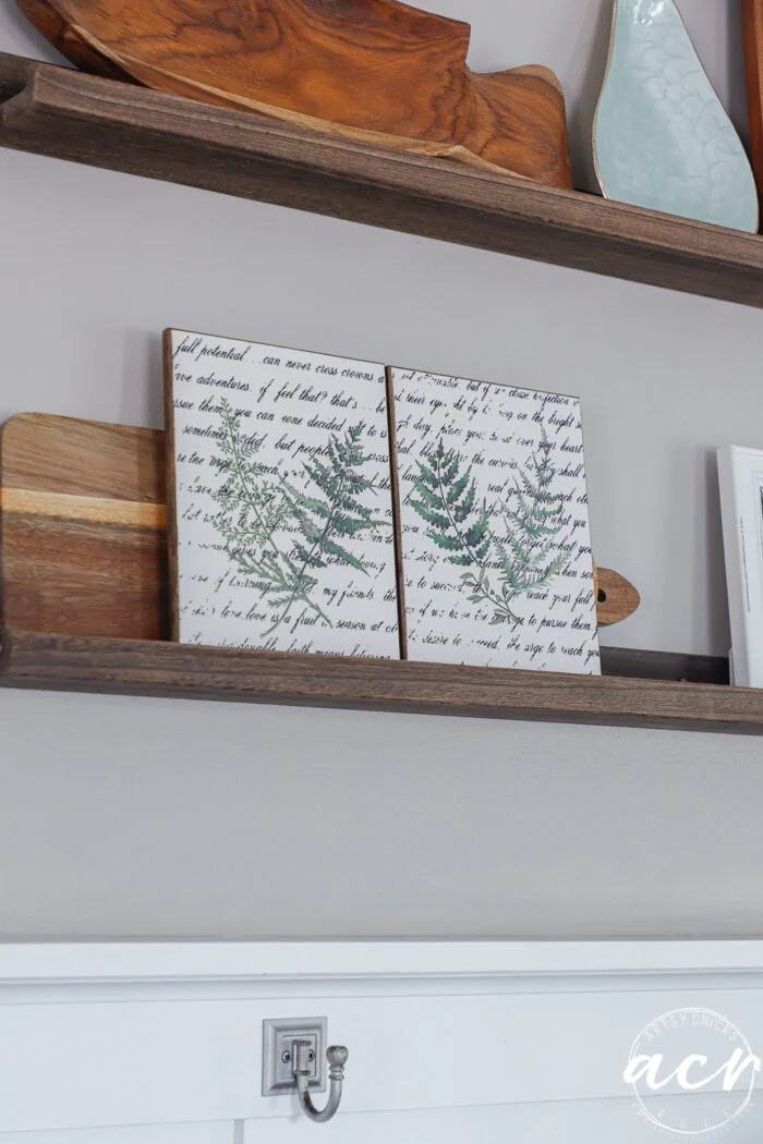 two wooden shelves display decorative items, including a green leaf-printed artwork on script background, wooden cutting boards, and ceramic pieces—all arranged for this easter edition of flipping smalls against a gray wall with a silver hook below.