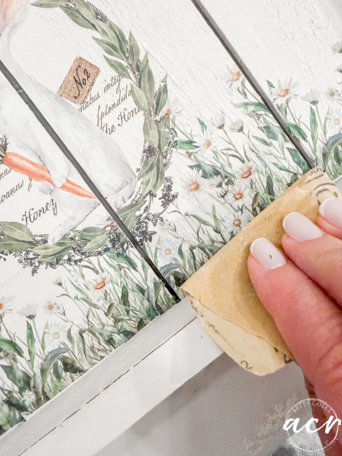 a hand with neatly manicured nails uses sandpaper to distress the edge of a painted wooden surface decorated with floral and greenery designs—perfect for your flipping smalls easter edition projects.