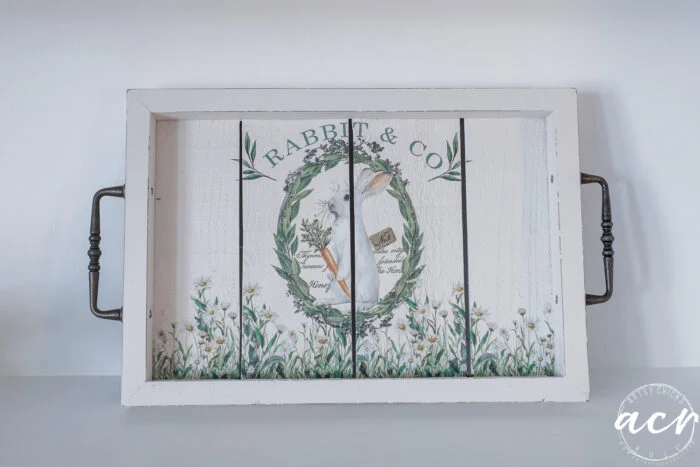 a white wooden tray with black handles features a floral wreath, carrots, and the text rabbit & co in green. part of the flipping smalls easter edition, it’s adorned with delicate white flowers and greenery along the edges.