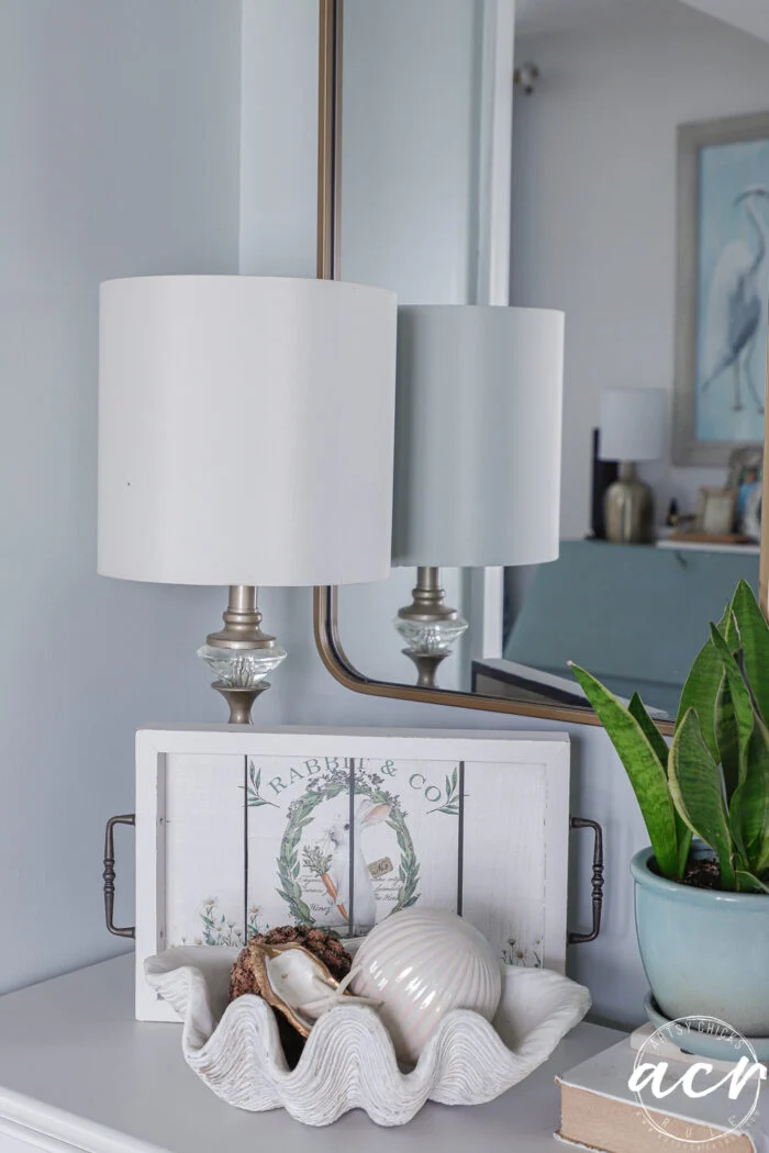 a white table holds a decorative clam shell with seashells, a potted plant, and a white lamp. behind the shell rests a white tray with floral art—an easter edition touch—while the mirror reflects this flipping smalls coastal-themed room.