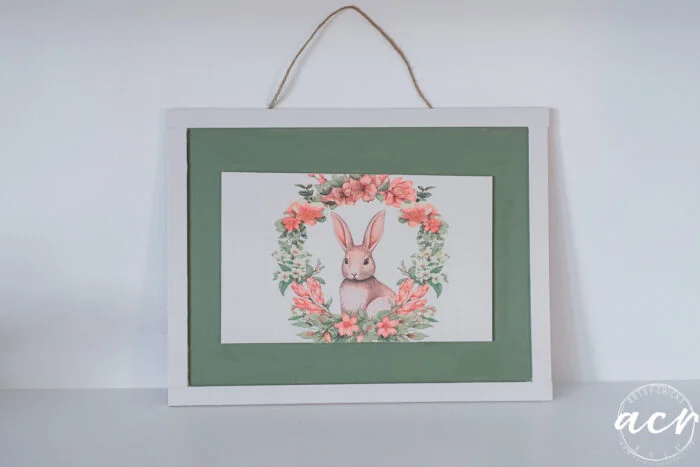 a framed picture of a brown rabbit surrounded by pink flowers and green leaves, hanging on a wall with a twine string. the frame is white with a green inner border, perfect for the flipping smalls easter edition collection.
