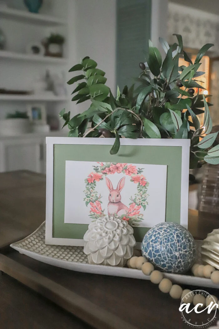a decorative tray on a table holds a framed bunny illustration with a floral wreath—perfect for an easter edition—alongside a potted leafy plant, textured balls, wooden bead garland, and a white carved artichoke ornament.