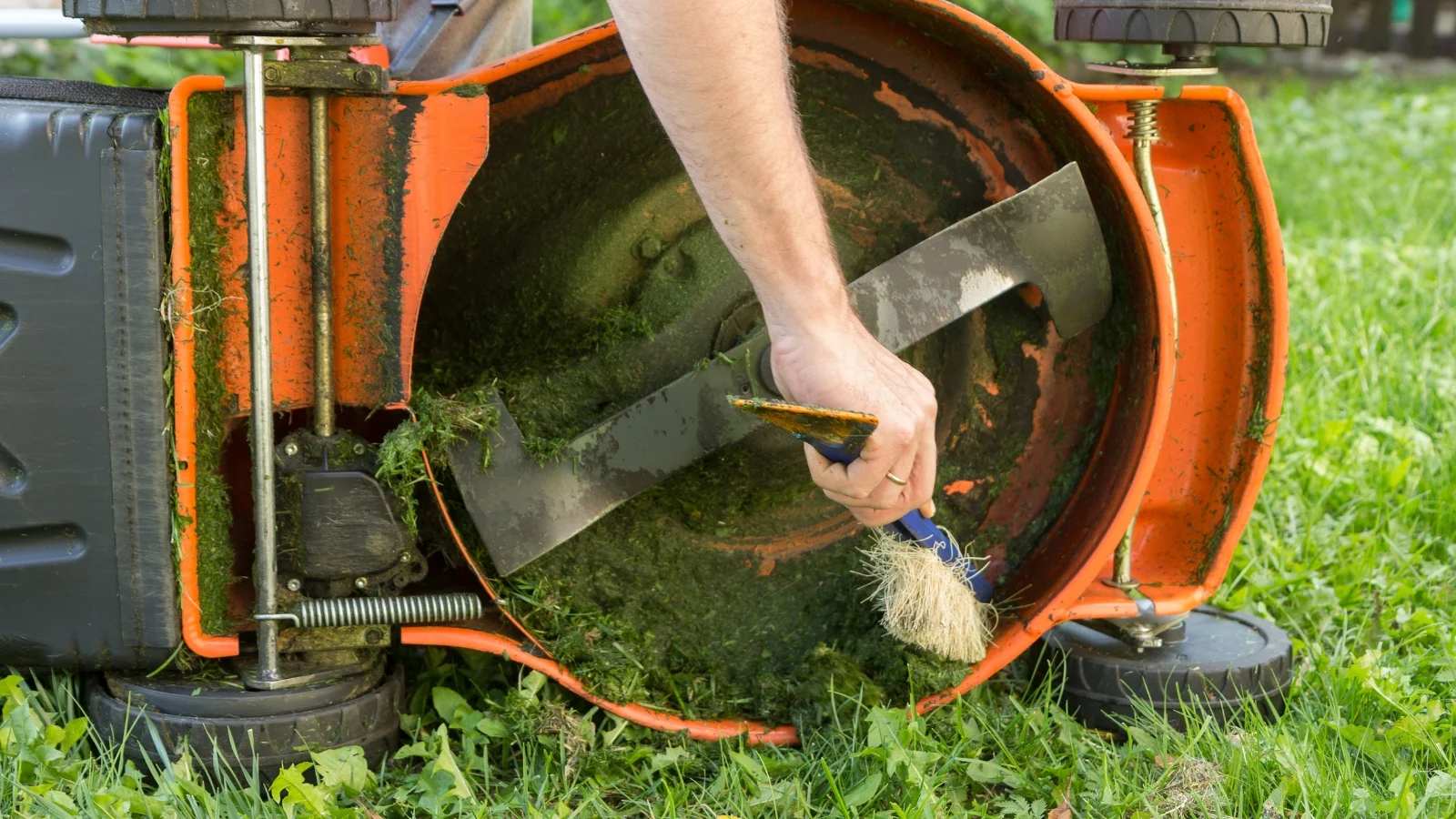 hands reaching under a mower to clear accumulated grass clippings and debris, focusing on the underside and blades to ensure clean operation.