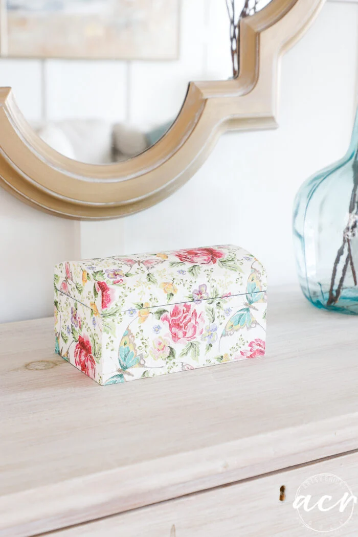 a small rectangular box with a domed lid, decorated using paper napkin decoupage in colorful floral and butterfly patterns, sits on a light wooden surface in front of a decorative mirror and a blue glass vase.