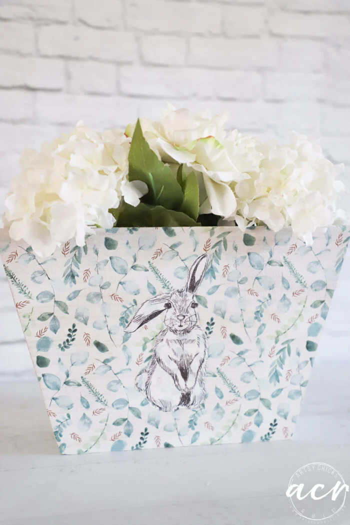 a floral arrangement with white flowers and green leaves in a decorative container featuring a rabbit drawing, perfect inspiration for learning how to decoupage with the iron on method, set against a white brick background.
