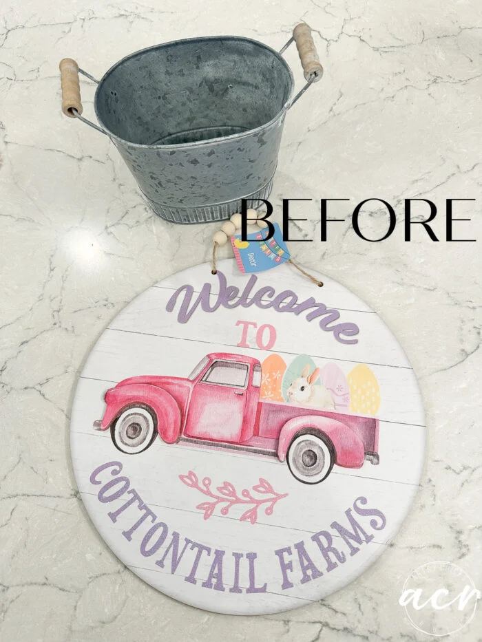 a round sign with a pink truck carrying easter eggs and a bunny, reading welcome to cottontail farms, lies on a marble surface next to an empty metal pail—perfect inspiration for napkin projects. the word before is overlaid on the image.