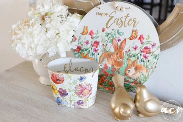 a spring-themed display inspired by napkin projects, featuring a floral bloom bucket, gold bunny figurines, white hydrangeas, and a decorative "happy easter" sign with rabbits and flowers.