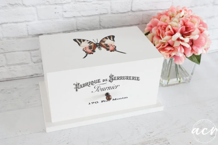 a white wooden butterfly card box with a butterfly illustration and french text sits on a white surface. pink hydrangea flowers in a glass vase are placed beside it, against a white brick wall backdrop.