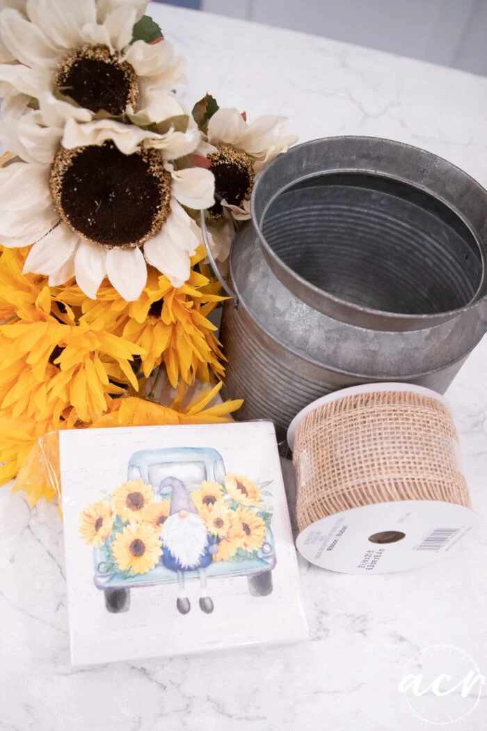 a bunch of artificial sunflowers, a galvanized metal bucket, a roll of burlap ribbon, and a decorative tile with a gnome in a truck filled with sunflowers create charming sunflower decor on a marble surface.