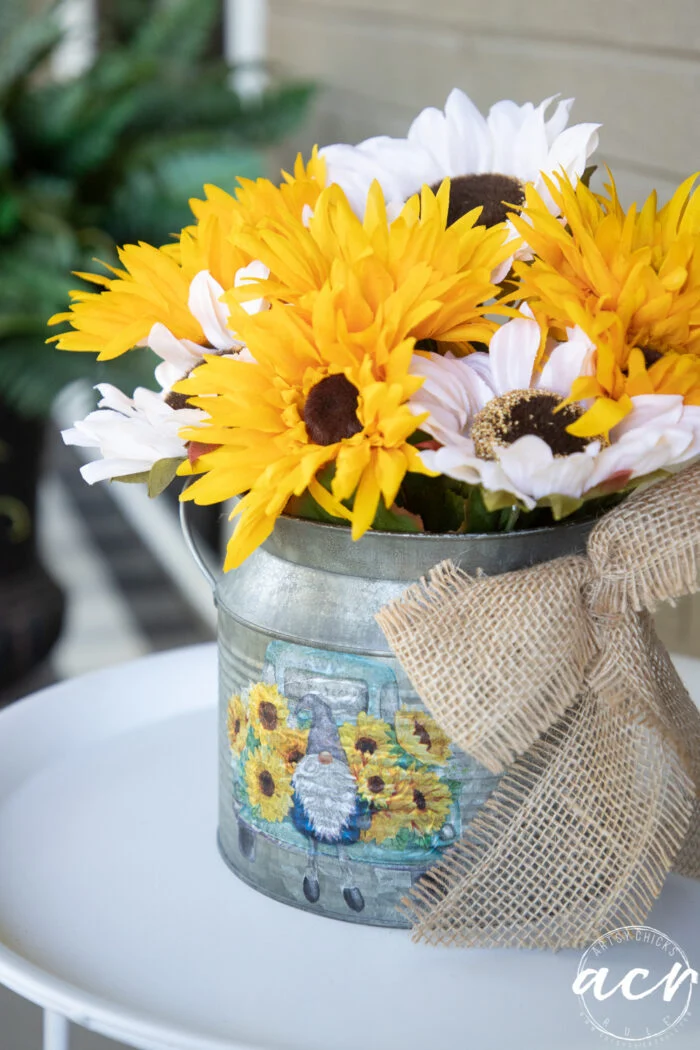 a charming metal watering can adorned with sunflower decor and a playful gnome is brimming with bright yellow sunflowers and white daisies, tied with a burlap bow, and sits gracefully on a white table.