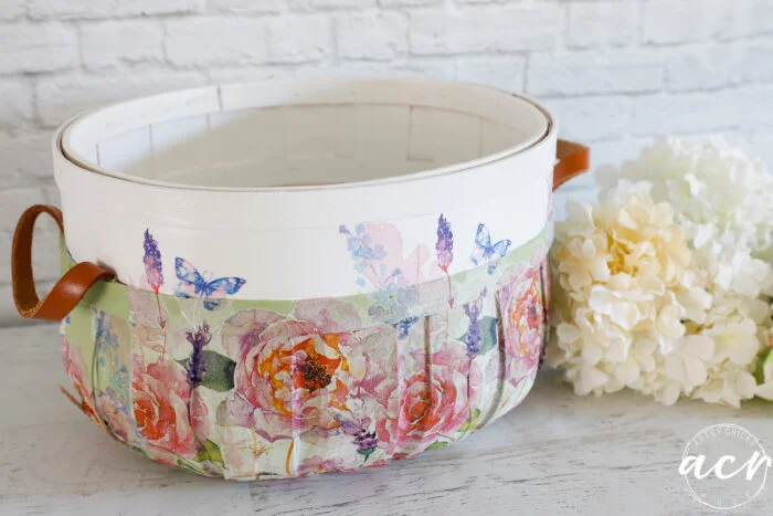 a white basket with leather handles features a colorful floral and butterfly design—an ideal basket update for spring. it sits on a white surface next to a bouquet of white hydrangeas, against a white brick background.