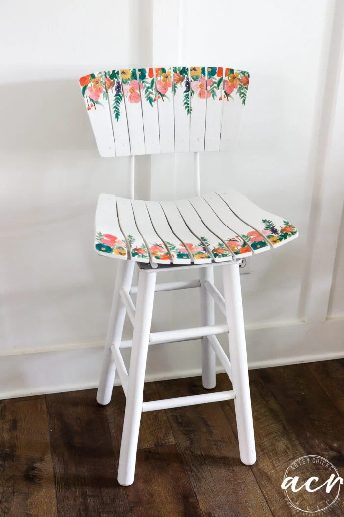 a white wooden chair with a slatted seat and backrest, featuring hand-painted colorful floral designs on the edges—a charming mid century modern slatted bar stool makeover. the chair sits on a dark wooden floor against a white paneled wall.