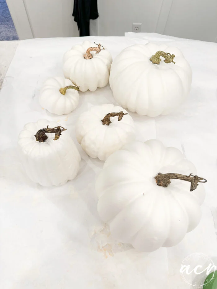 six white decoupage pumpkins with napkins of varying sizes, each with brown stems, are arranged on a white surface. the minimal background draws focus to the unique pumpkins.