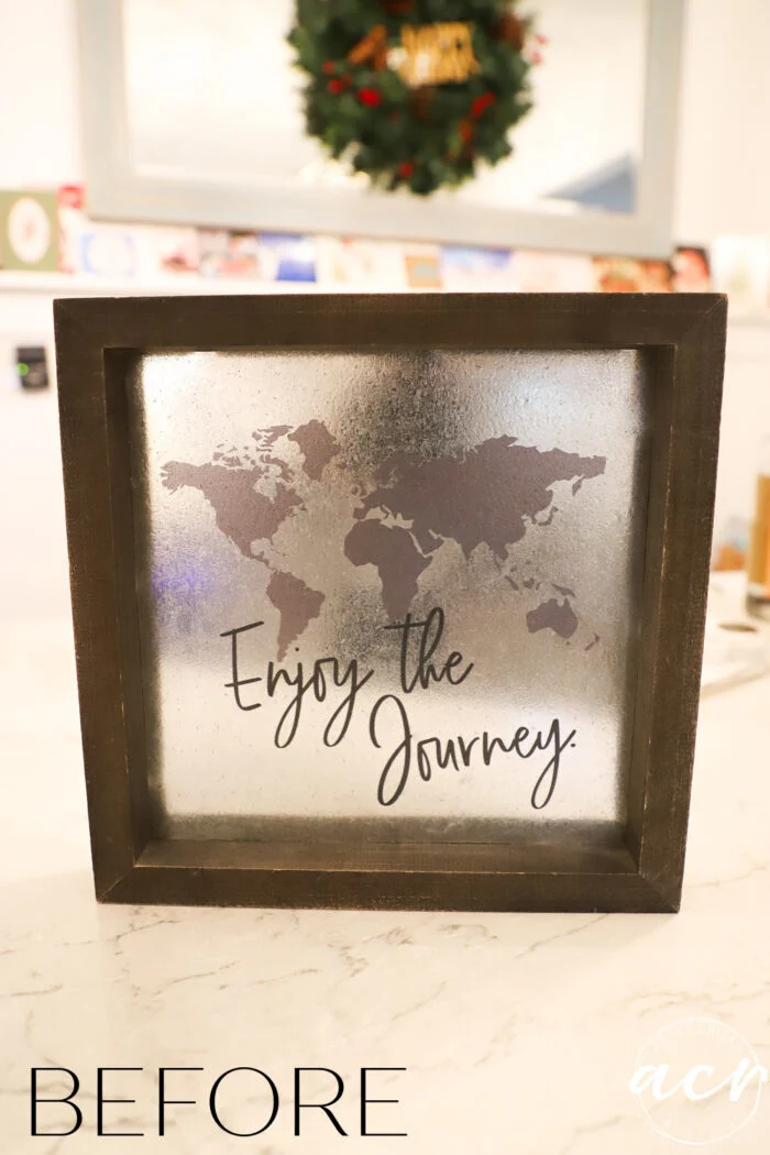 a framed sign with a world map silhouette and the words “enjoy the journey” shines on a reflective metallic background, sitting on a marble countertop. the text before appears at the bottom left, complementing an and to all a good night sign decor style.