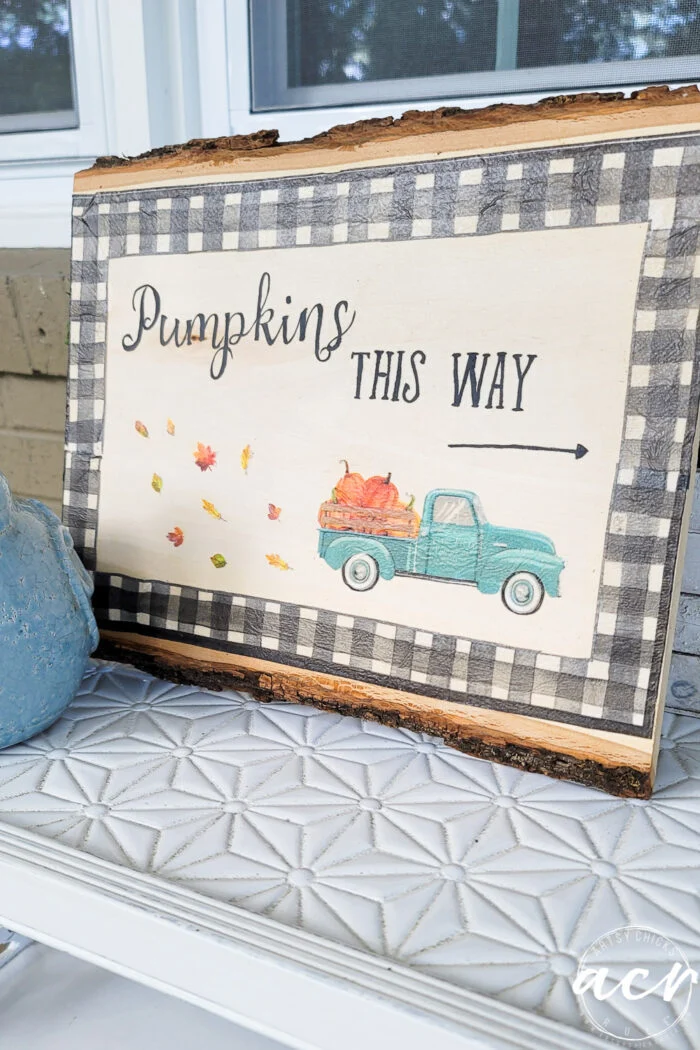 a wooden sign with a black-and-white plaid border features napkin decoupage details, reading pumpkins this way with an arrow, falling leaves, and a teal vintage truck carrying pumpkins, propped on a white patterned surface.