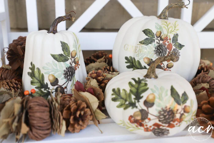 three white decoupage pumpkins with napkins, featuring botanical illustrations of leaves, acorns, and pinecones, are surrounded by pinecones and autumn foliage for a festive fall display on a white surface.