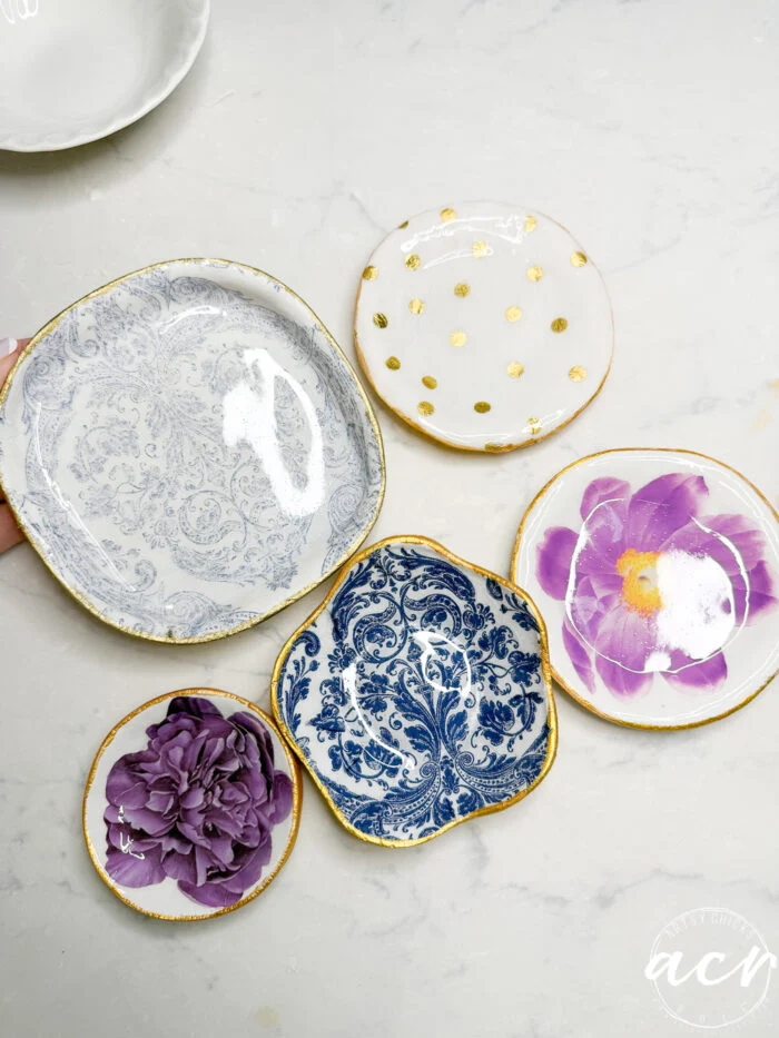 five small, decorative ceramic plates with gold edges—perfect for diy air dry clay projects—are arranged on a white surface. designs include blue patterns, gold polka dots, and purple flowers. a hand holds one of the plates.