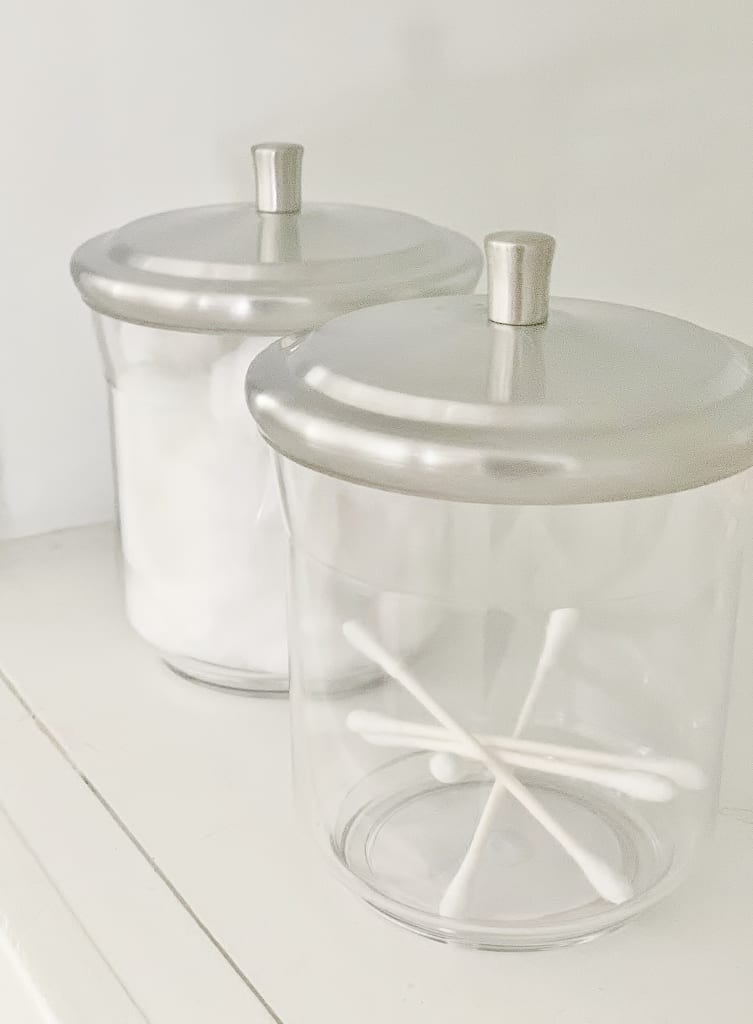 two glass jars with silver lids, sitting on a white shelf in the midst of a hallway makeover. the front jar contains cotton swabs, visible through its clear glass. the second jar appears similar but