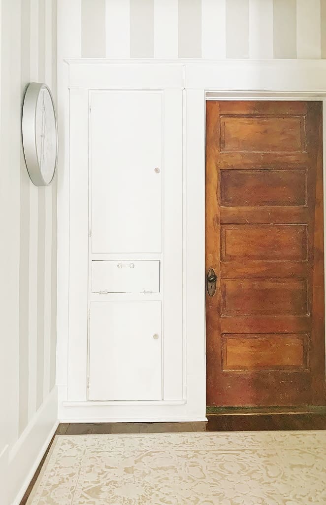 a hallway makeover featuring a white storage cabinet next to a wooden door, with a round wall mirror mounted to the left, set against a striped wallpaper background and patterned floor.
