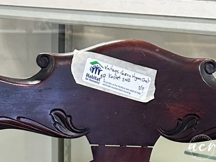 a close-up of a wooden chair with a habitat for humanity price tag that reads, vintage chair cherry hyman co. basket seat, placed on the decorative backrest.