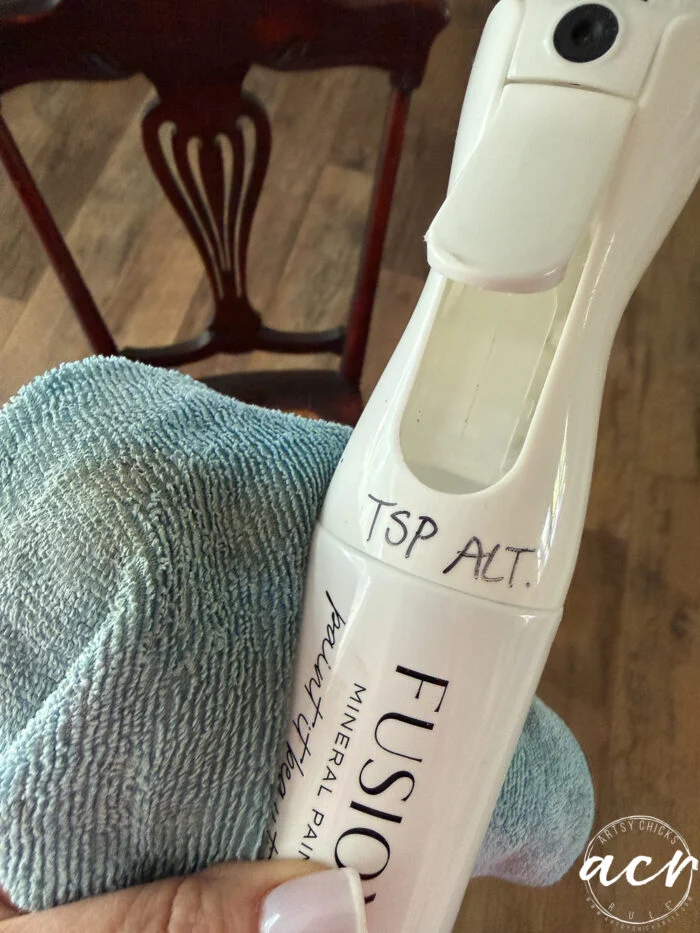 a hand holds a spray bottle labeled tsp alt. and a blue cleaning cloth, preparing for a chair update, with a vintage chair and wooden floor in the background.